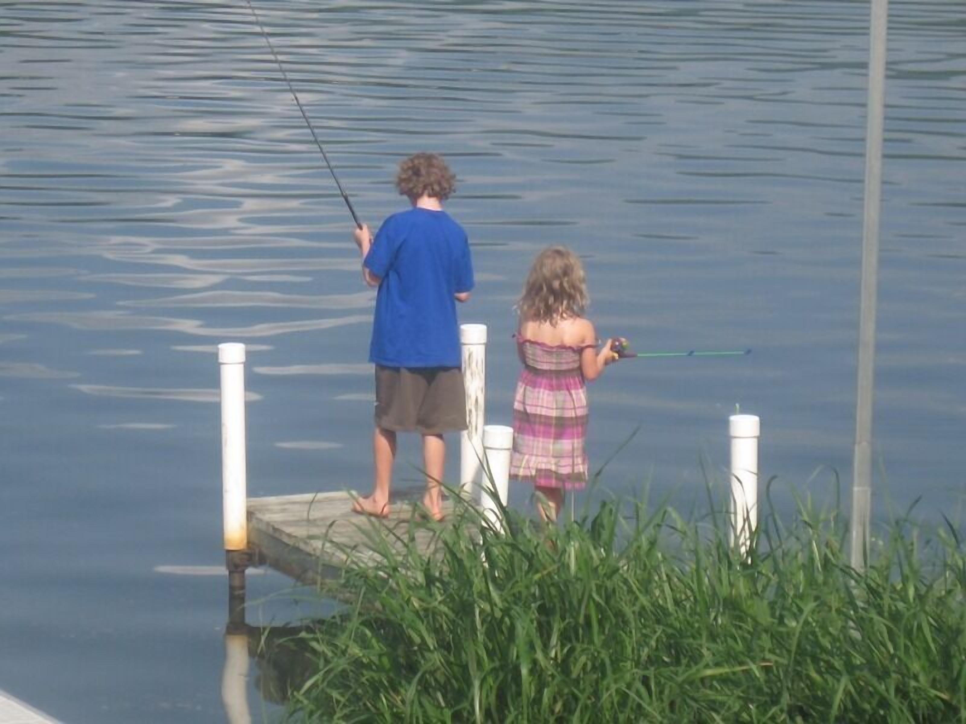 Two people fishing on a dock by a calm lake.