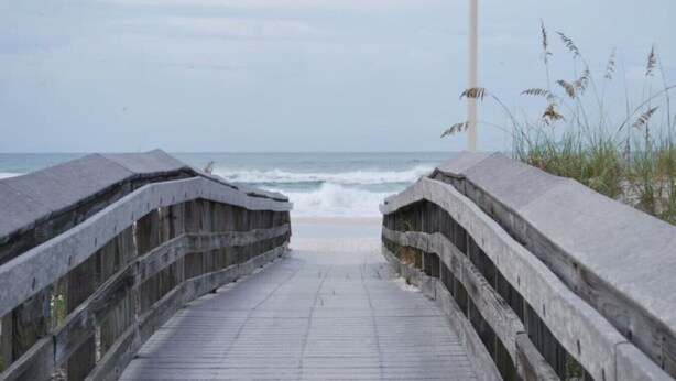 En støbt træpromenade der fører til stranden med udsigt over havet.