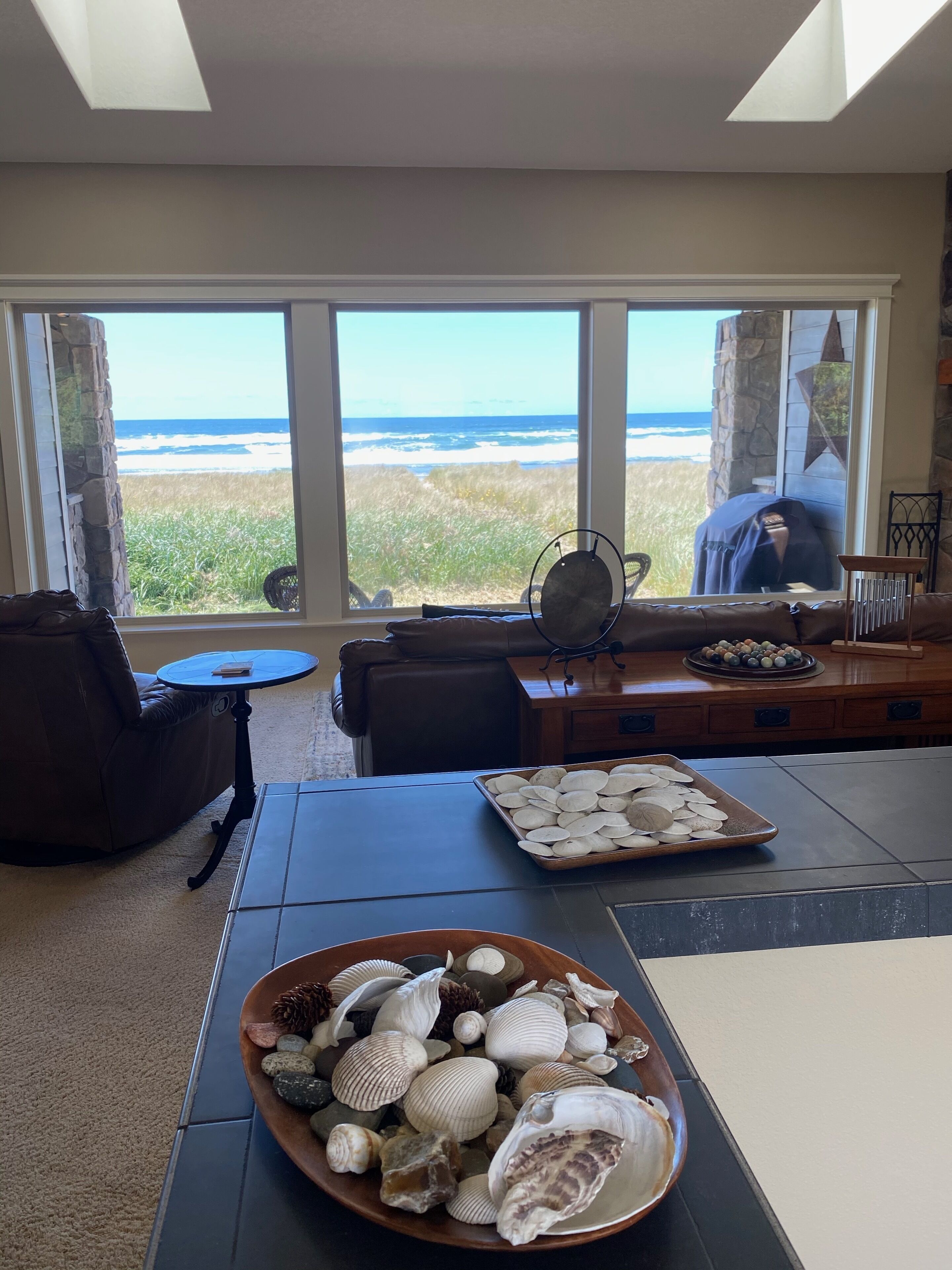 A room with a large window overlooking the ocean, a stone wall, a leather sofa, a small round table, and a tray of seashells.