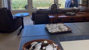 A room with a large window overlooking the ocean, a stone wall, a leather sofa, a small round table, and a tray of seashells. - Beach Star - Classic, Elegant & Comfortable Beachfront Home! (Rockaway Beach)