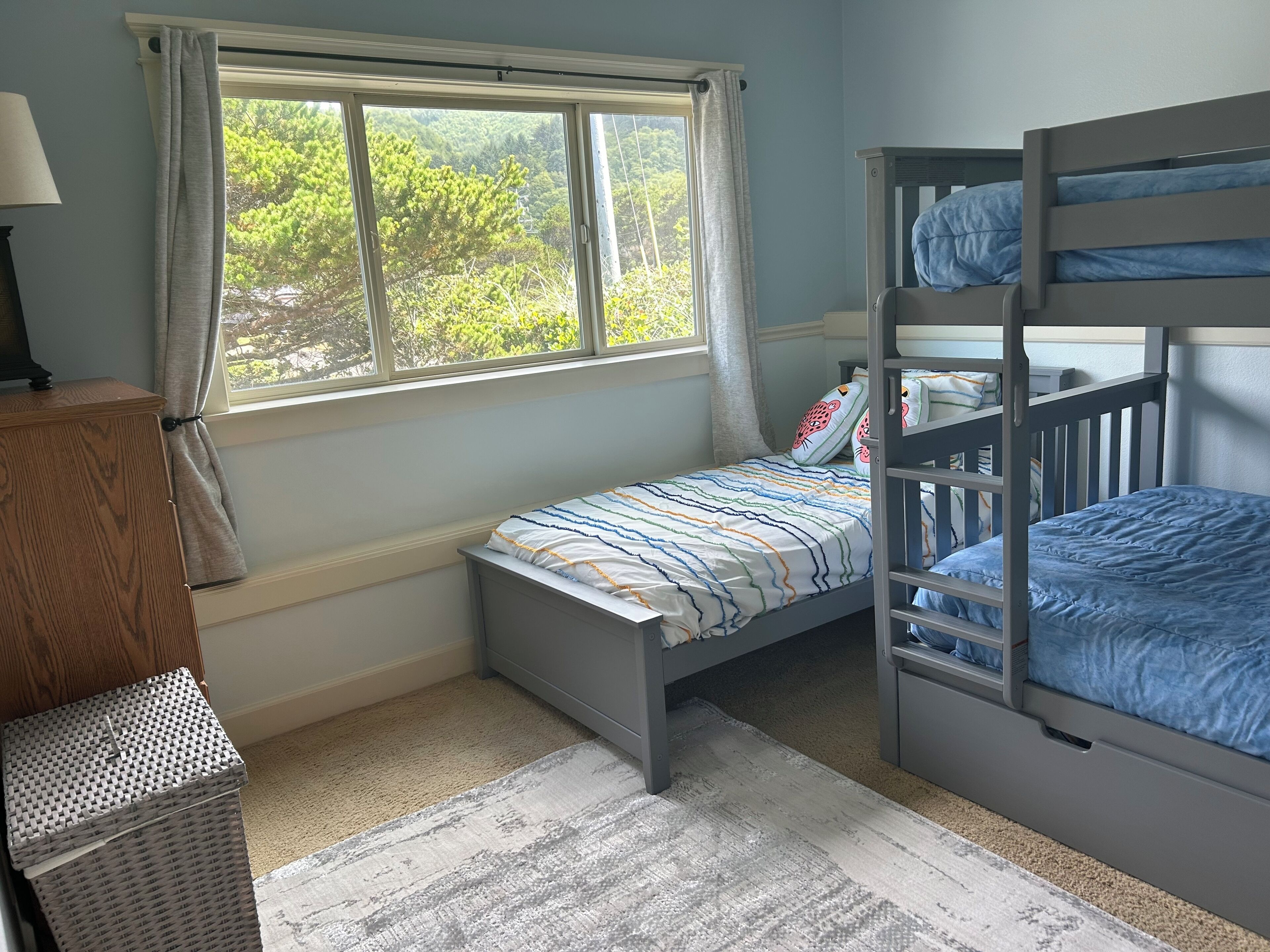 A bedroom with a bunk bed, a single bed, a window with curtains, a lamp, and a dresser.