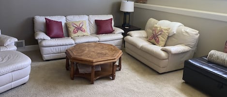 Un salon doté d’un tapis beige, d’un canapé blanc agrémenté de coussins rouges et beiges, d’une table basse en bois et d’une fenêtre donnant sur un paysage verdoyant.