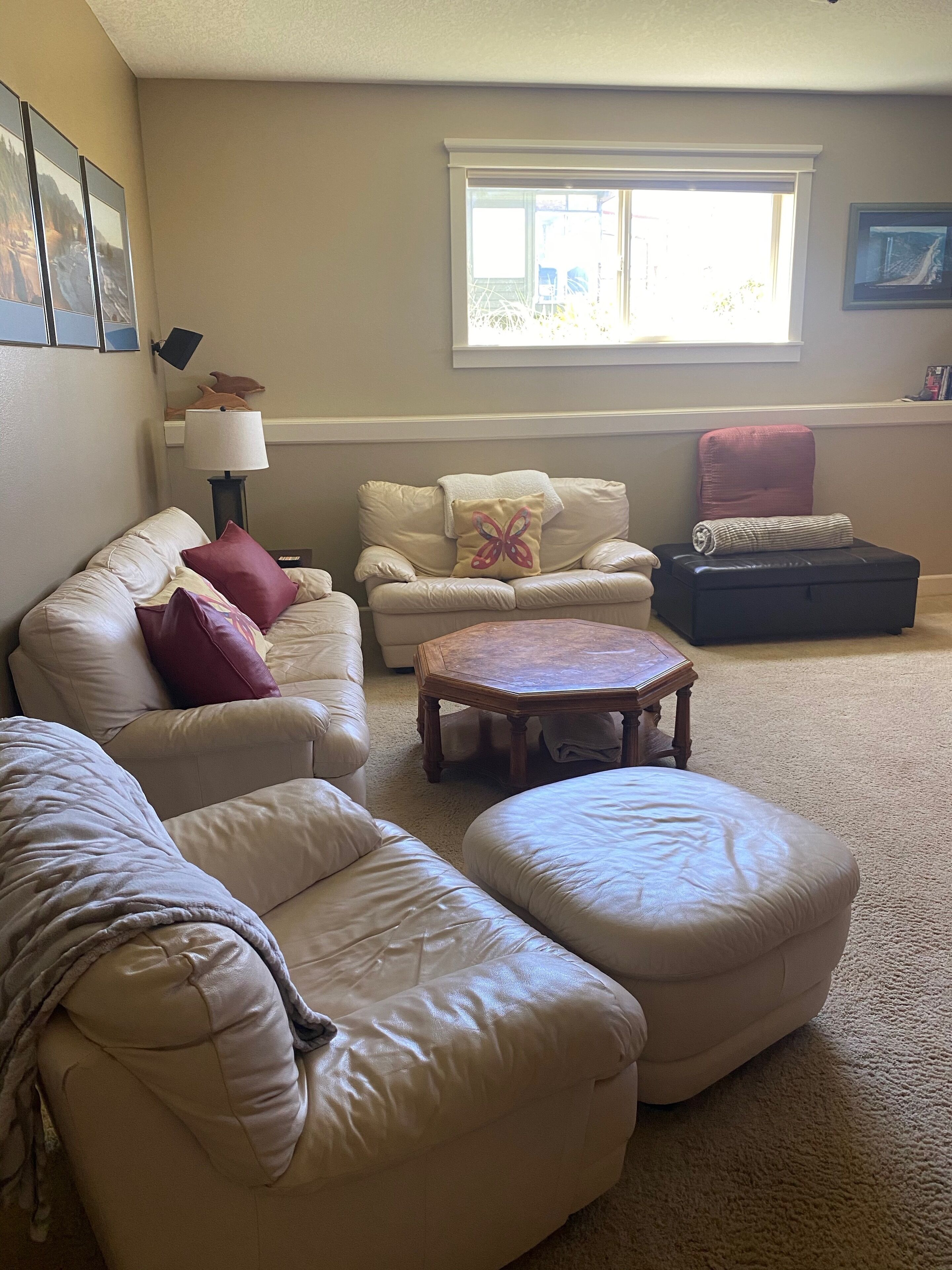 A living room with a beige carpet, a beige sofa, a matching ottoman, a wooden coffee table, and a window with blinds.