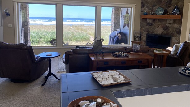 A living room with a large window overlooking the beach, a stone fireplace, and a dining area with a tray of seashells. - Beach Star - Classic, Elegant & Comfortable Beachfront Home! (Rockaway Beach)