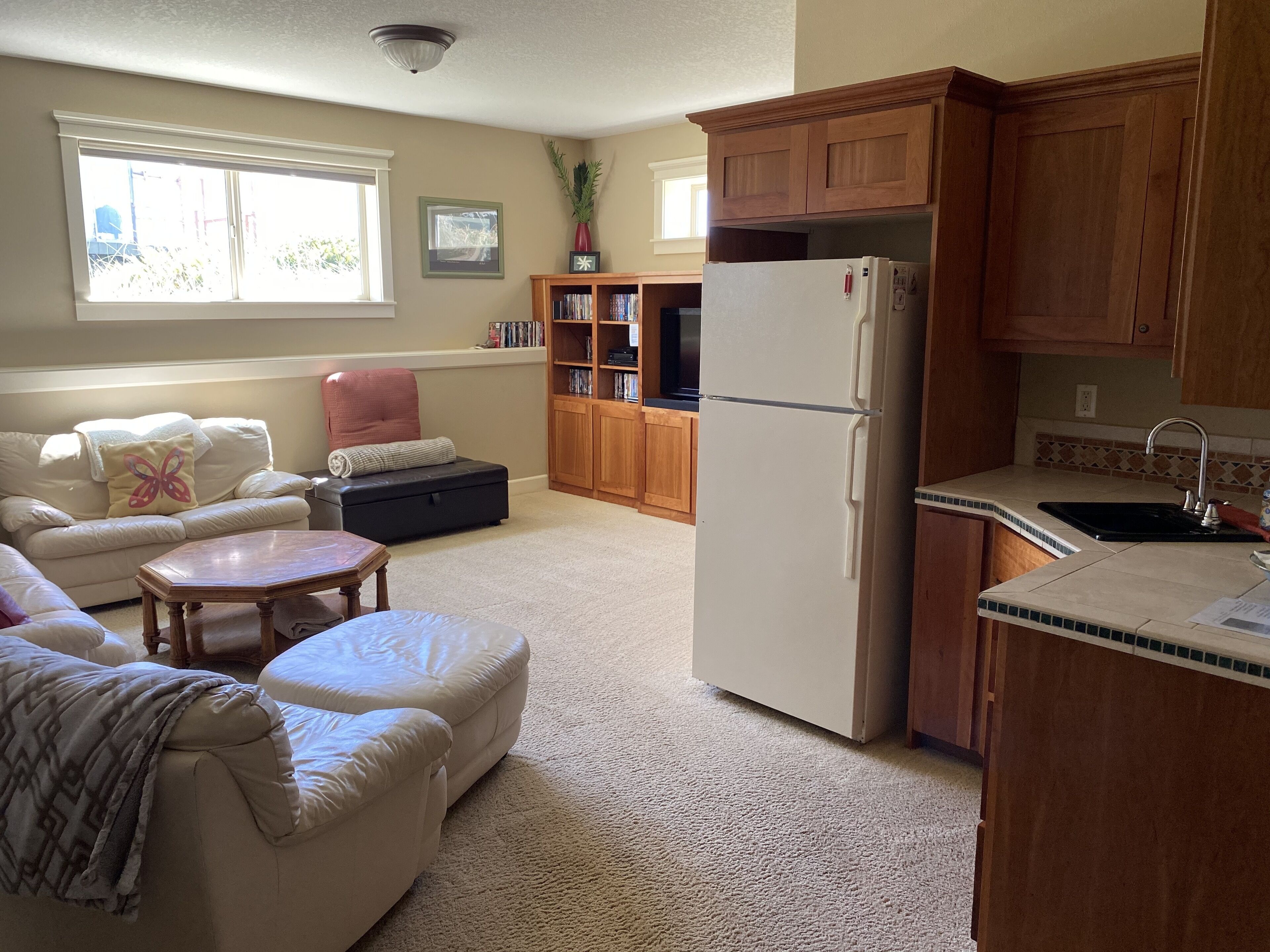 A living room with a sofa, ottoman, and coffee table, a kitchen with wooden cabinets and a refrigerator, and a bookshelf with books and decorative items.