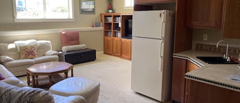 A living room with a sofa, ottoman, and coffee table, a kitchen with wooden cabinets and a refrigerator, and a bookshelf with books and decorative items.