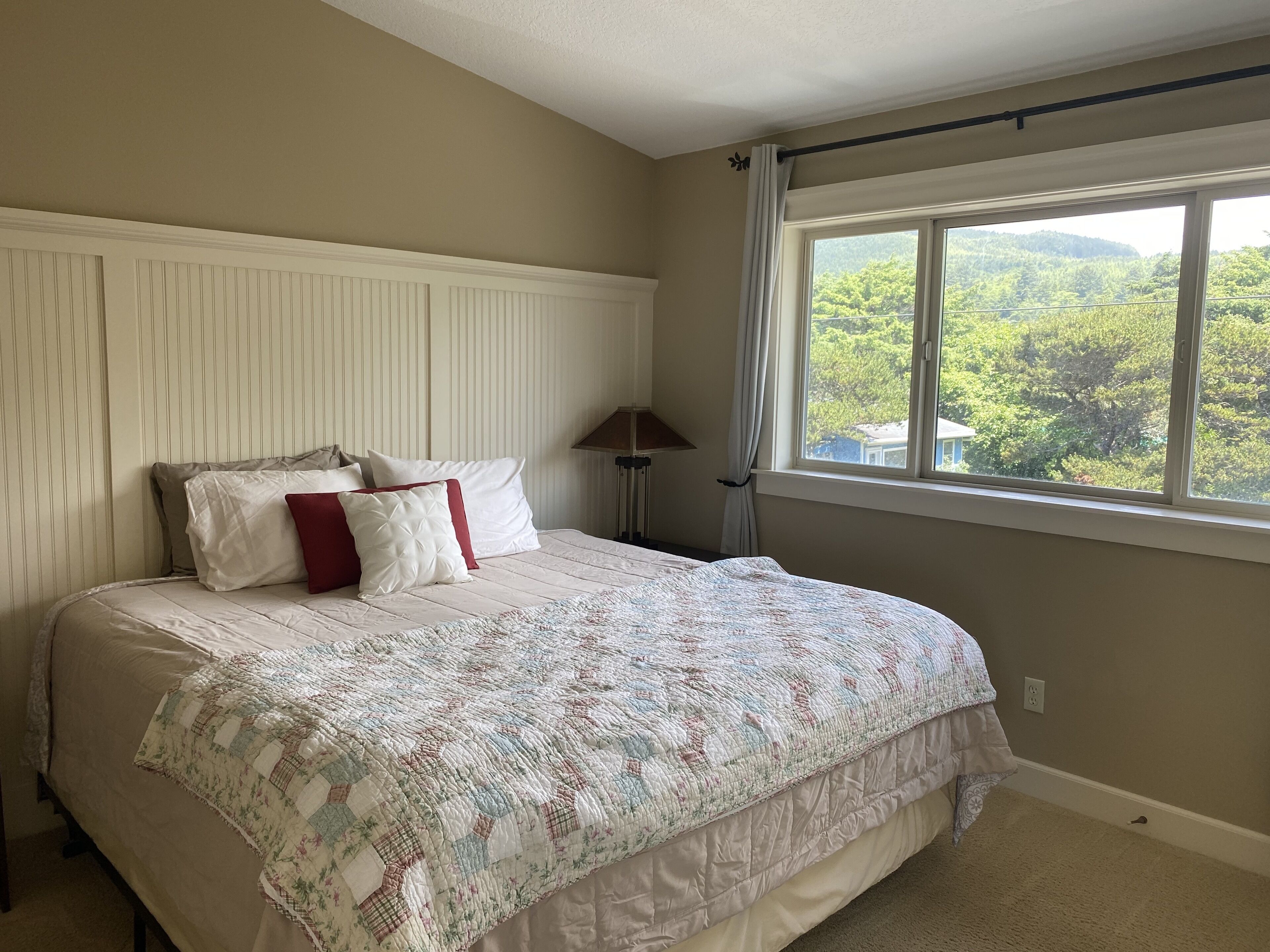 A bedroom with a large bed, a window with a view of greenery, and a bedside lamp.