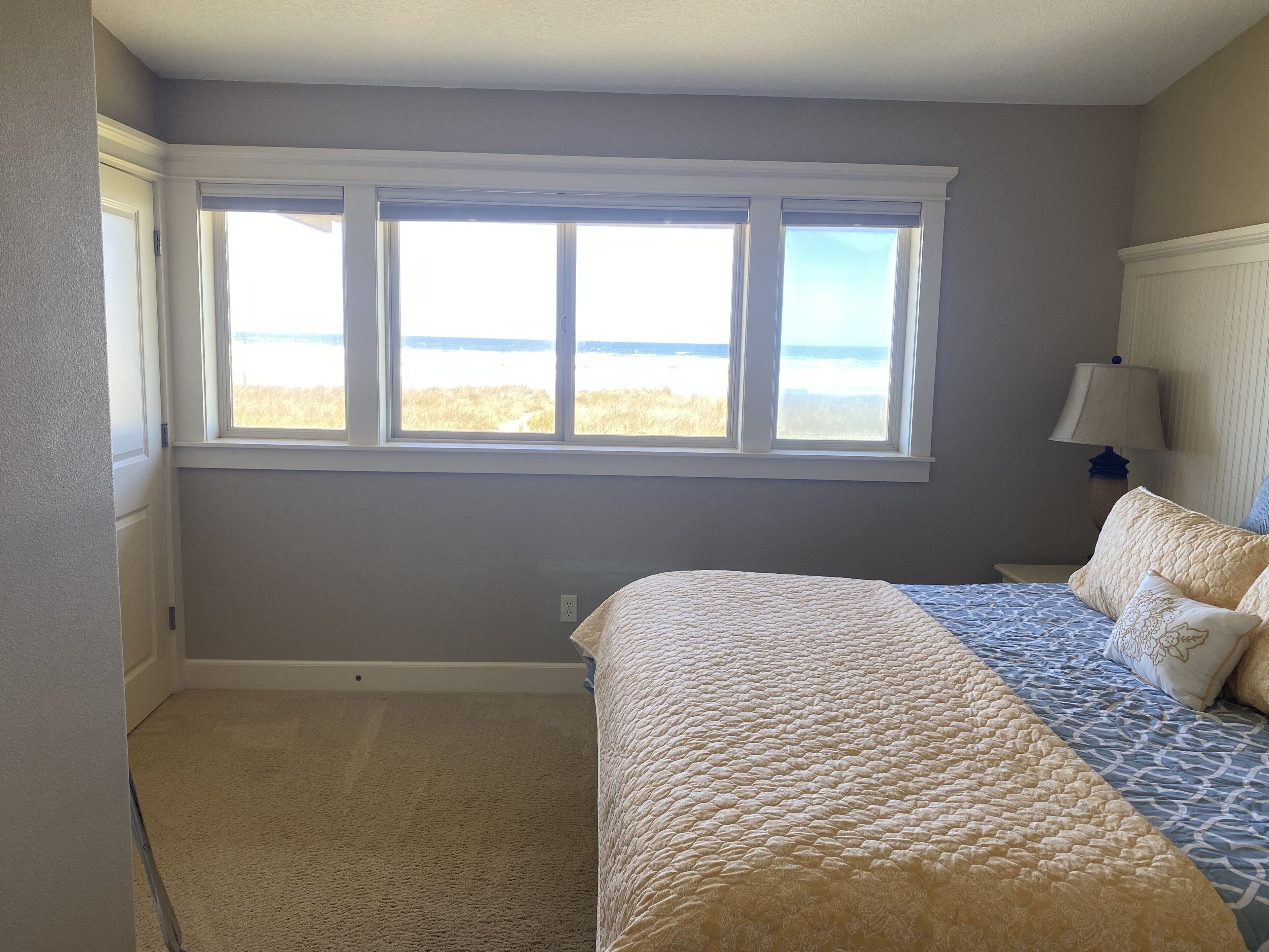 A bedroom with a large window, a bed with patterned bedding, and a nightstand with a lamp.