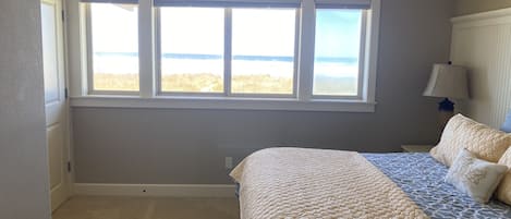 A bedroom with a large window, a bed with patterned bedding, and a nightstand with a lamp.