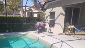 A swimming pool with clear water, a covered grill, a patio area with a table and chairs, and a house with a tiled roof.