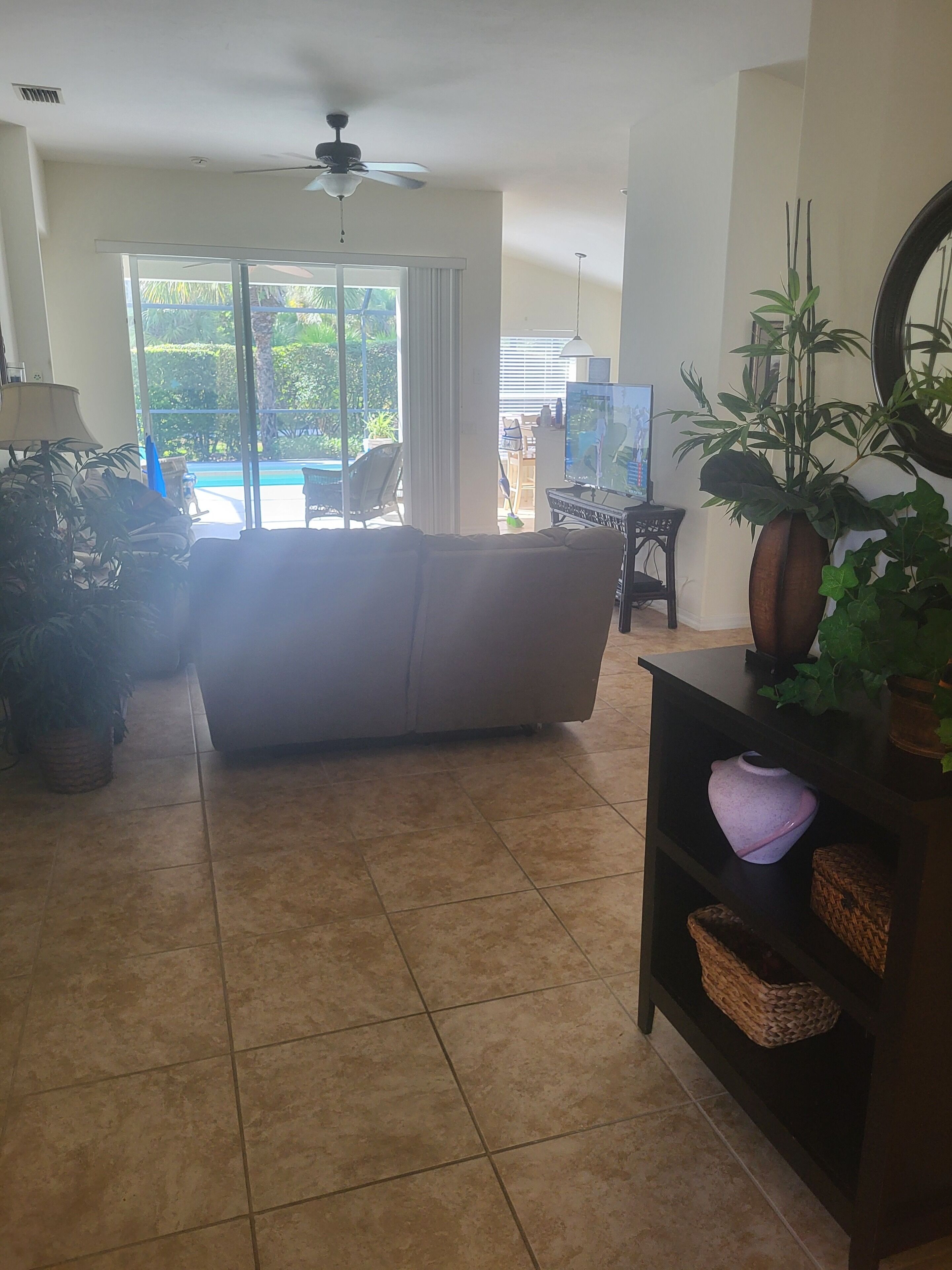 A living room with a sofa, a TV, a ceiling fan, and a large window with a view of a pool and greenery.
