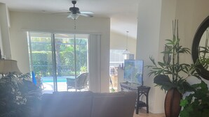 A living room with a sofa, a TV, a ceiling fan, and a large window with a view of a pool and greenery.