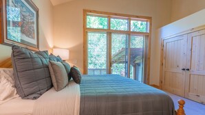 Une chambre à coucher avec un grand lit, une tête de lit en bois, une fenêtre avec des stores, un tableau encadré au mur et une porte avec un panneau en bois.