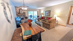 A dining area with a wooden table, chairs, and a vase of flowers. There is a sofa and a lamp in the living area.