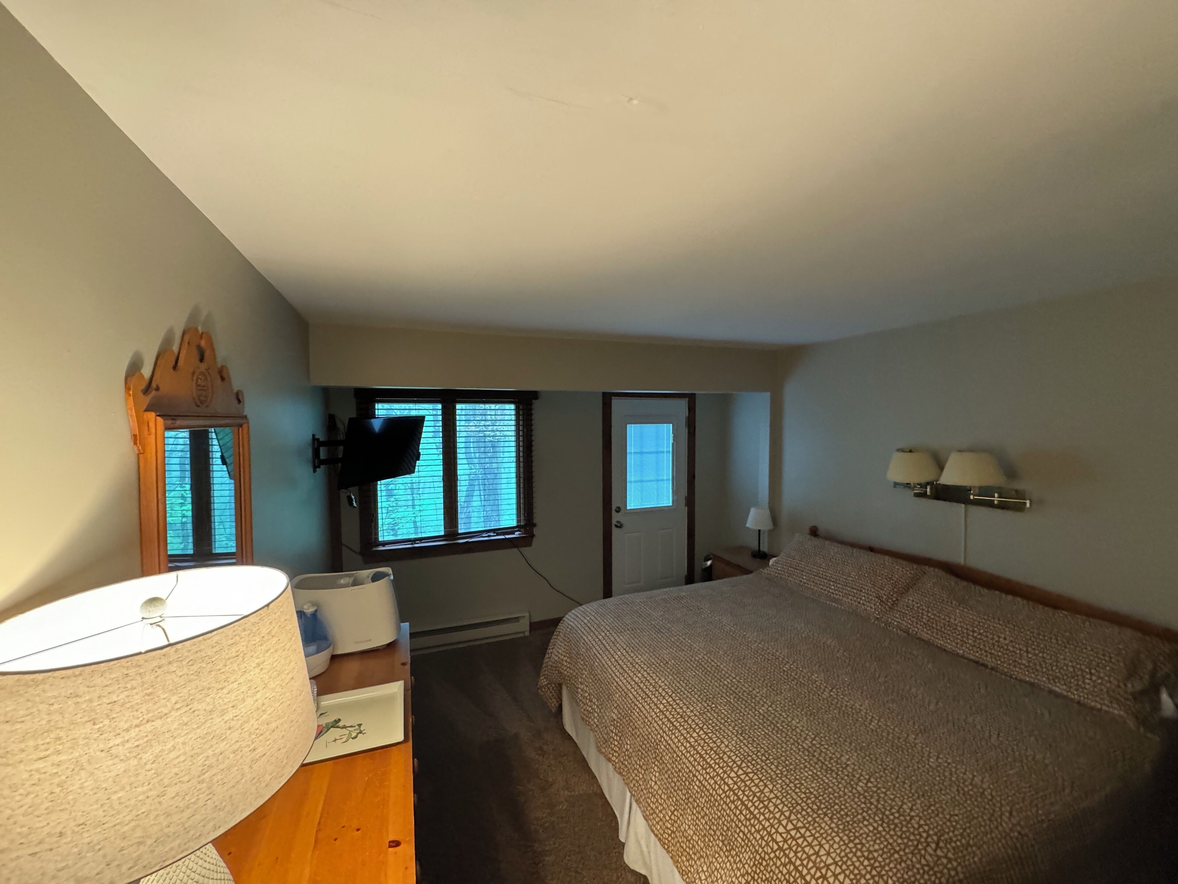 A bedroom with a bed, a desk with a lamp, a window, and a door.
