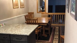 Una cocina con encimera de granito, armarios de madera, comedor con mesa y sillas de madera, una lámpara de araña y una alfombra estampada.