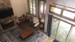 Una sala de estar con chimenea de piedra, un sofá gris, una mesa de centro de madera y un ventanal con persianas.