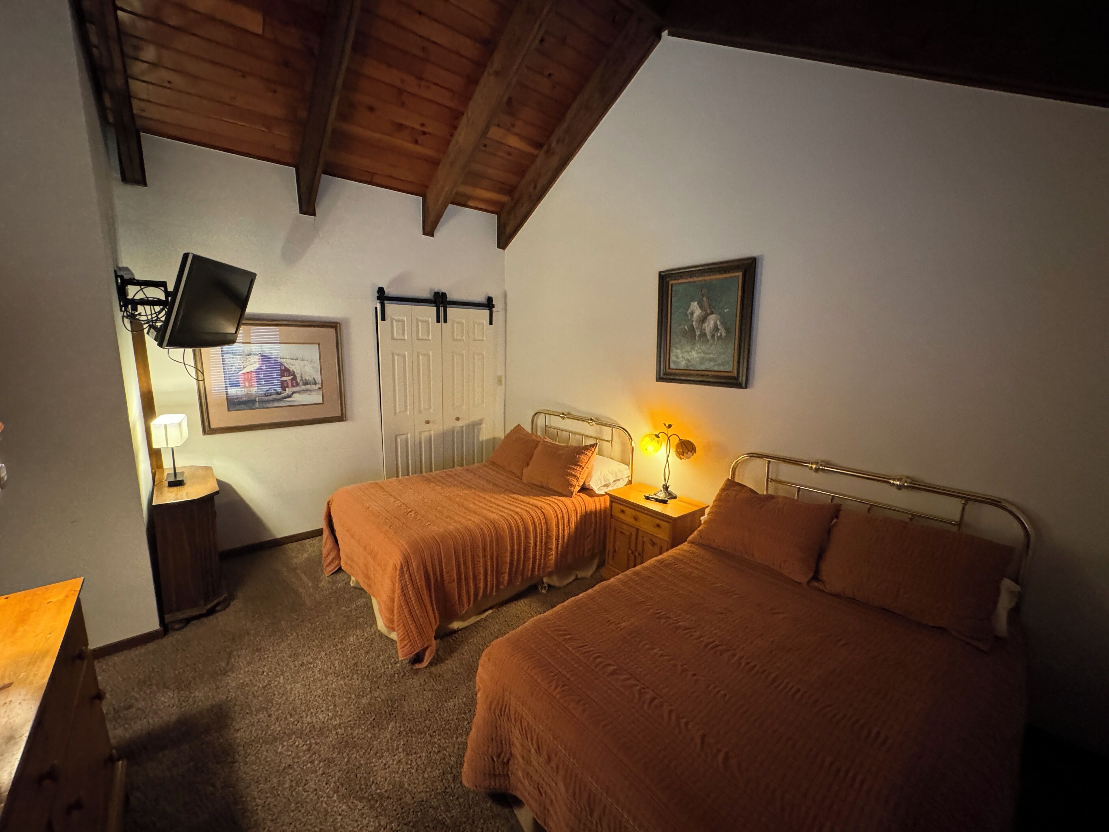 Un dormitorio con dos camas, un televisor montado en la pared, un cuadro enmarcado y una mesita de noche con una lámpara.