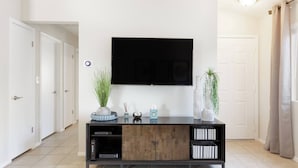A modern living room with a flat-screen TV mounted on the wall, a wooden console table with decorative items, and a tiled floor. - The "Pool Cottage" Upgraded Home FREE Heated Pool (Phoenix)