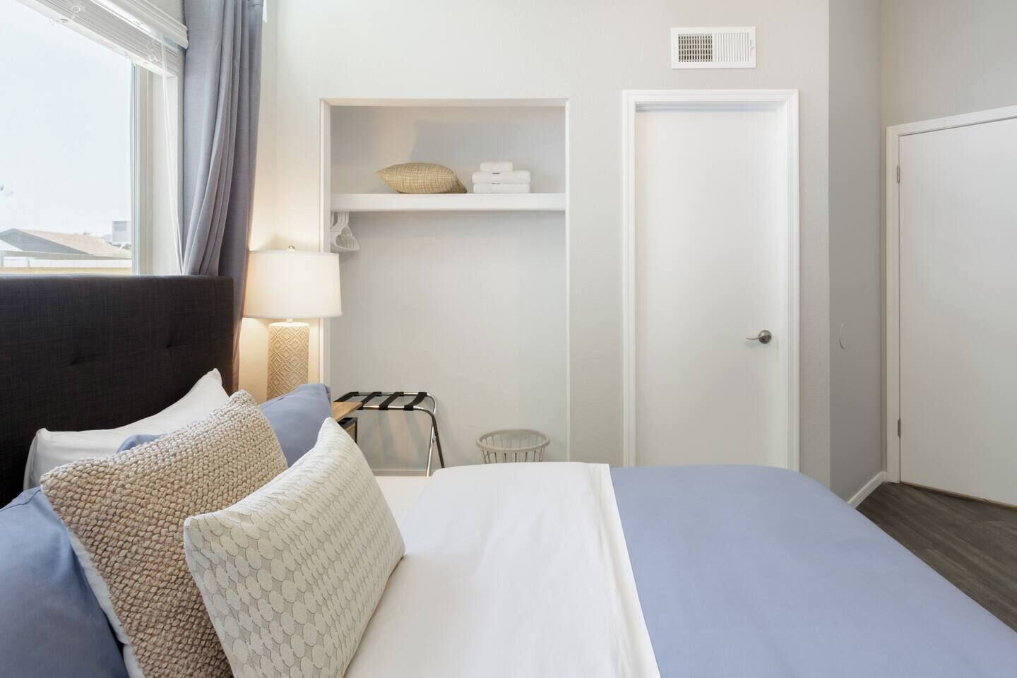 A hotel room with a bed, pillows, a bedside table with a lamp, a wardrobe, and a shelf with towels and a basket.