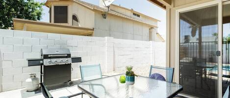 An outdoor patio with a grill, a table with chairs, and a view of a house in the background.