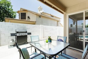 Une terrasse extérieure avec un barbecue, une table et des chaises, et une vue sur une maison en arrière-plan.