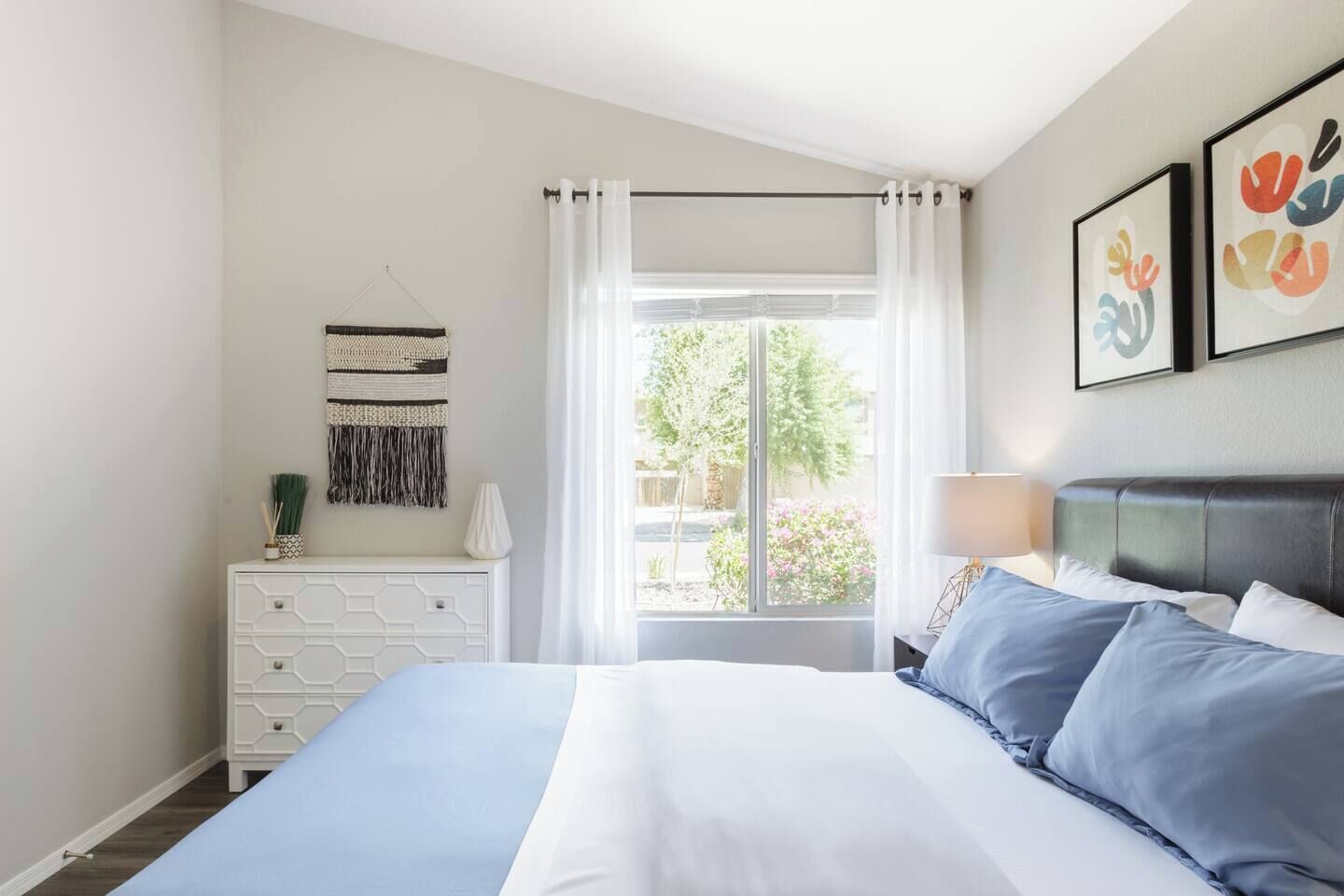 Une chambre à coucher comprenant un lit, une commode, une fenêtre avec des rideaux et deux œuvres d’art encadrées accrochées au mur.