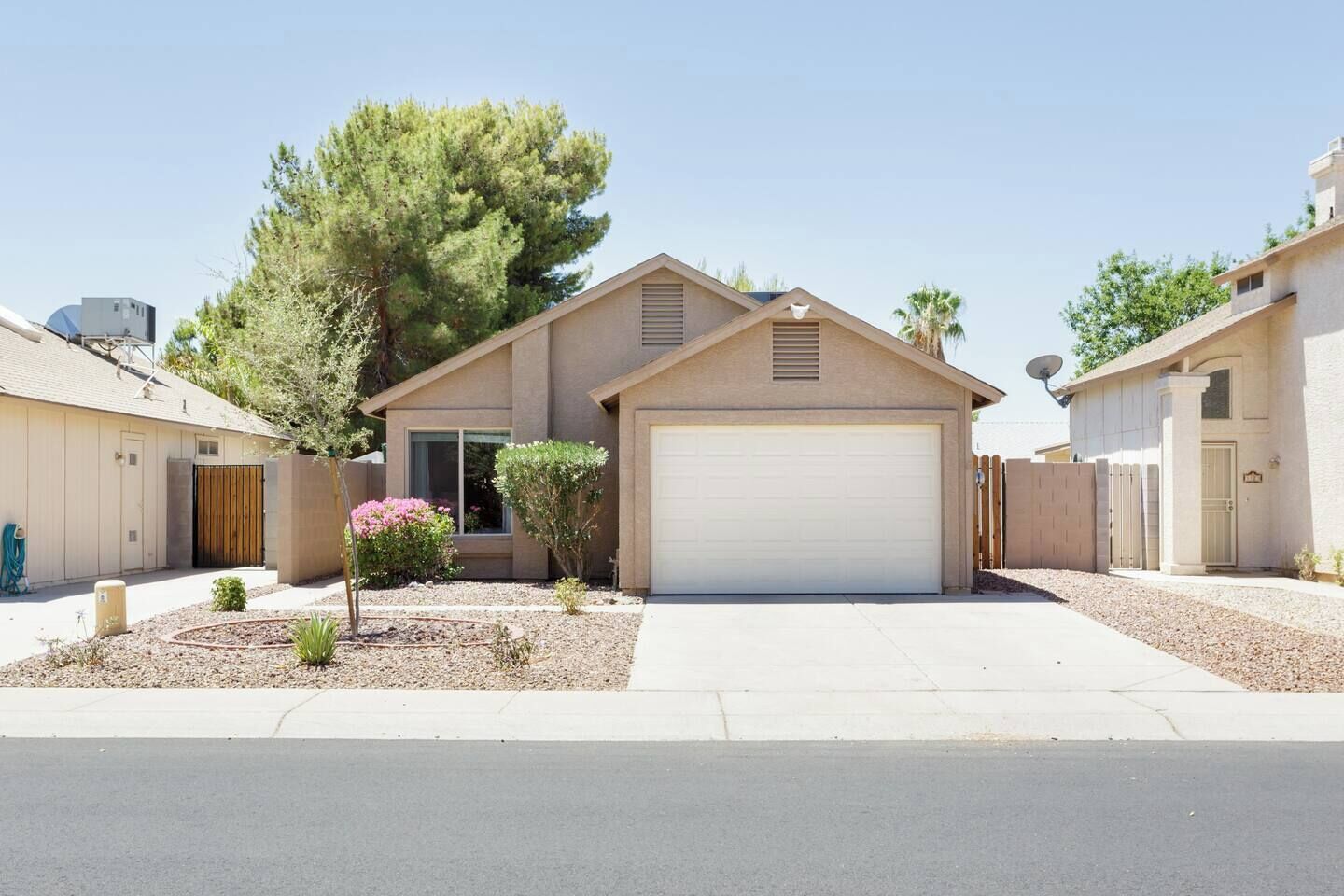 A single-story house with a garage, a small front yard with a tree and shrubs, and a clear sky.