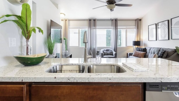 A modern kitchen with a granite countertop, stainless steel sink, and a dishwasher. There is a ceiling fan, a large window with curtains, and a dining area with a sofa and table. - The "Pool Cottage" Upgraded Home FREE Heated Pool (Phoenix)