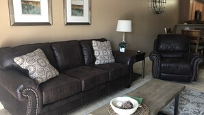 A living room with a dark brown leather sofa, a matching armchair, a coffee table with a woven placemat, and two framed landscape paintings on the wall. - Great location!! Pool! Unit 5201- Mtn. View Condos, 3 Bed, 2 Bath (Pigeon Forge)