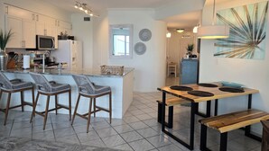Eine moderne Küche mit weißen Schränken, einer zentralen Kochinsel mit Barhocker, einem Essbereich mit Holztisch und Bänken sowie einem großen abstrakten Gemälde an der Wand.