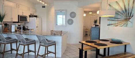 Una cocina moderna con armarios blancos, una isla central con taburetes de bar, una zona de comedor con mesa de madera y bancos, y un cuadro abstracto grande en la pared.