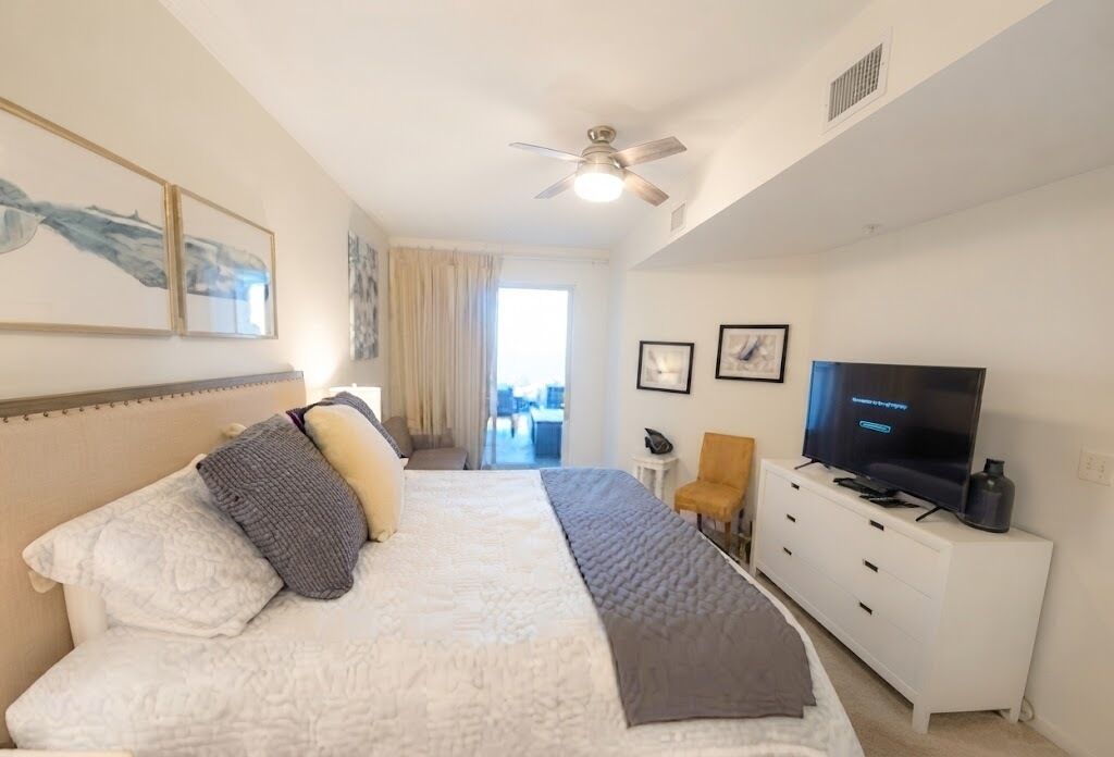 A hotel room with a bed, a TV, a ceiling fan, and artwork on the walls.
