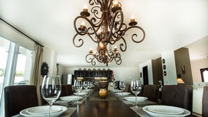 A dining room with a dark wooden table set for six, surrounded by brown leather chairs. A large, ornate chandelier hangs from the ceiling, and a vase with flowers is placed in the center of the table.