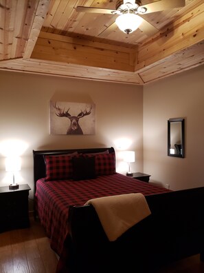 A wooden-paneled room with a bed, two bedside lamps, a ceiling fan, and a wall-mounted mirror. - Luxury Rustic Estate on Lake Freeman (Monticello)