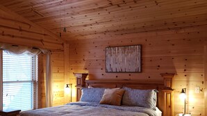 Une chambre dans une cabane en bois, avec un lit, des tables de chevet, un ventilateur de plafond et une fenêtre avec des rideaux.