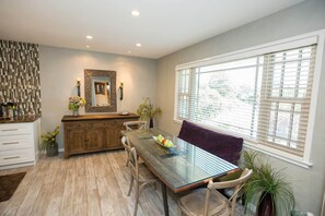 A dining area with a glass table, chairs, a wooden cabinet, and a large window with blinds.