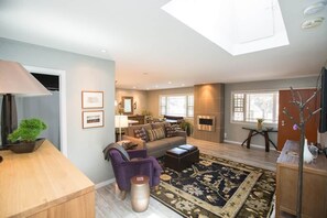 Una sala de estar moderna con sofá, sillón, mesa de centro y un tapete grande. Hay un comedor con mesa y sillas, una zona de cocina y un tragaluz.