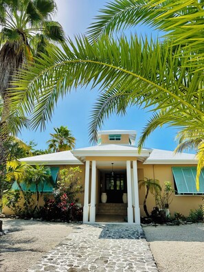 A house with a stone pathway leading to a white porch, surrounded by palm trees and tropical plants. - You Know you want to be Here!/Sleeps 6/Private Pool and Fun Tiki Bar! (Providenciales)
