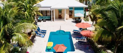 Un espace piscine doté de chaises longues et de parasols, entouré de palmiers et bordé d’un bâtiment au toit blanc.