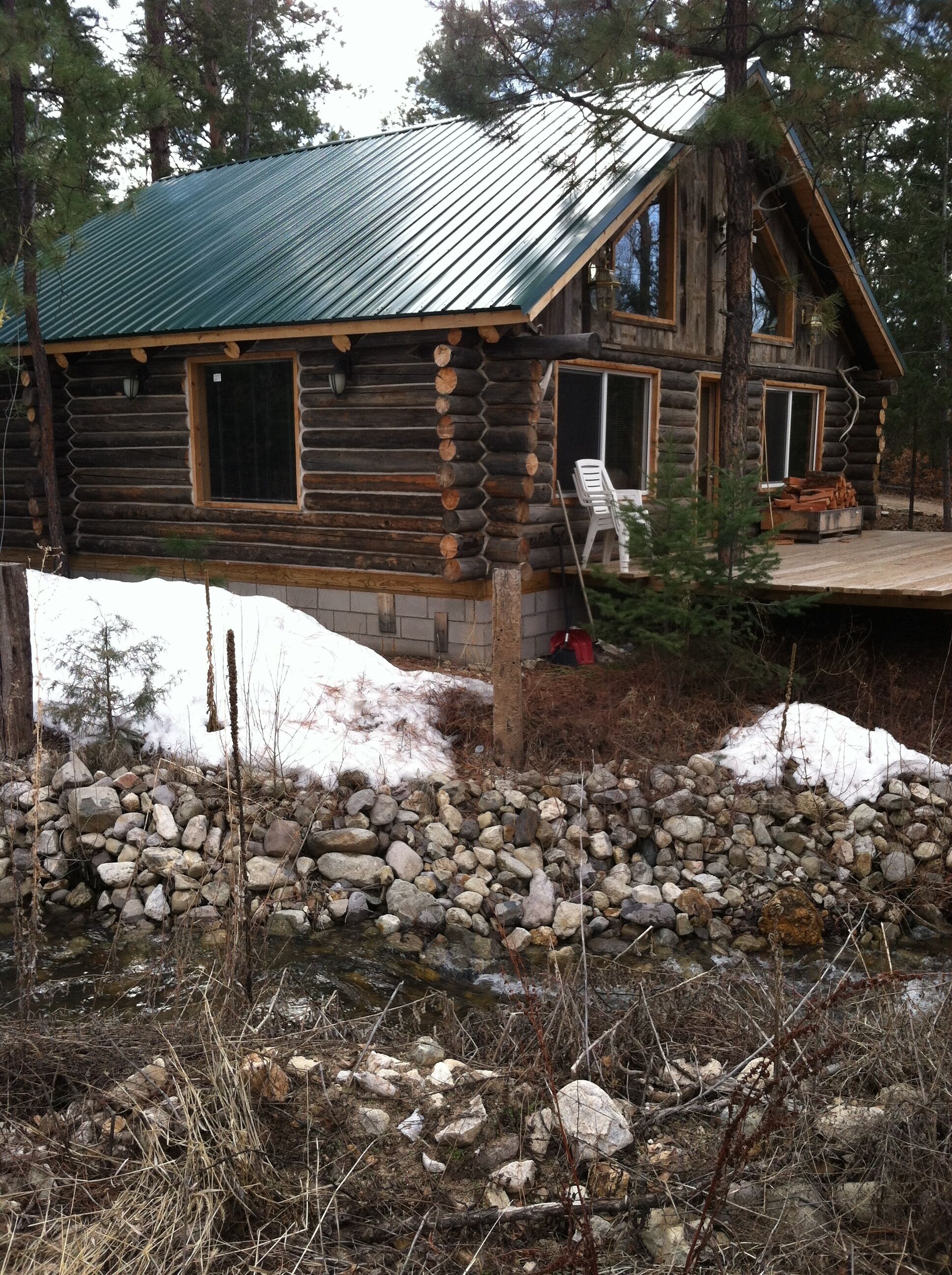 A log cabin with a metal roof, surrounded by a stone wall and snow patches.