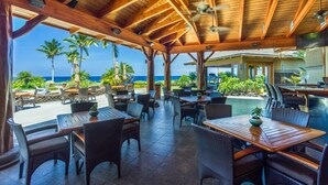 Un espace extérieur aménagé en terrasse, avec des tables et des chaises en bois, donnant sur une piscine et des palmiers, et offrant une vue sur l’océan.