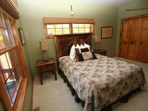A bedroom with a large bed, a wooden headboard, a window with blinds, a nightstand with a lamp, and a wooden wardrobe.