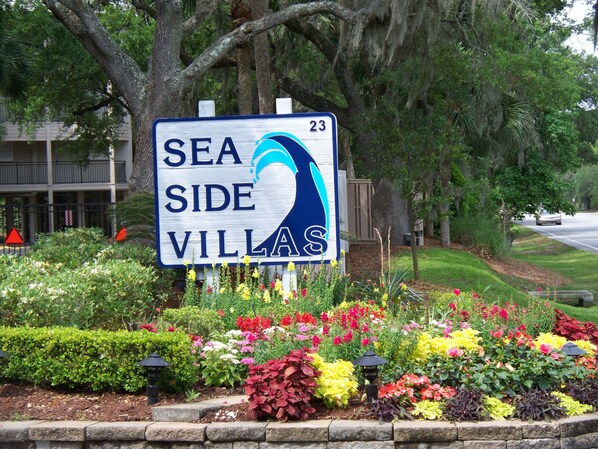 A sign for Sea Side Villas with a wave design, surrounded by colorful flower beds. - AWESOME REMODEL! FREE BIKES, ALL BEACH GEAR, on the beach, 5-star reviews. (hilton head island)