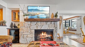 A cozy living room with a stone fireplace, a large flat-screen TV mounted above it, and a comfortable seating area with patterned cushions.