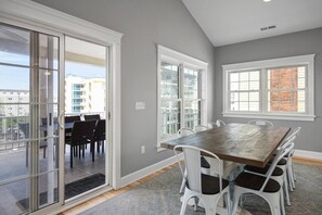 A dining area with a wooden table and white chairs, large windows with white frames, and a view of buildings outside.