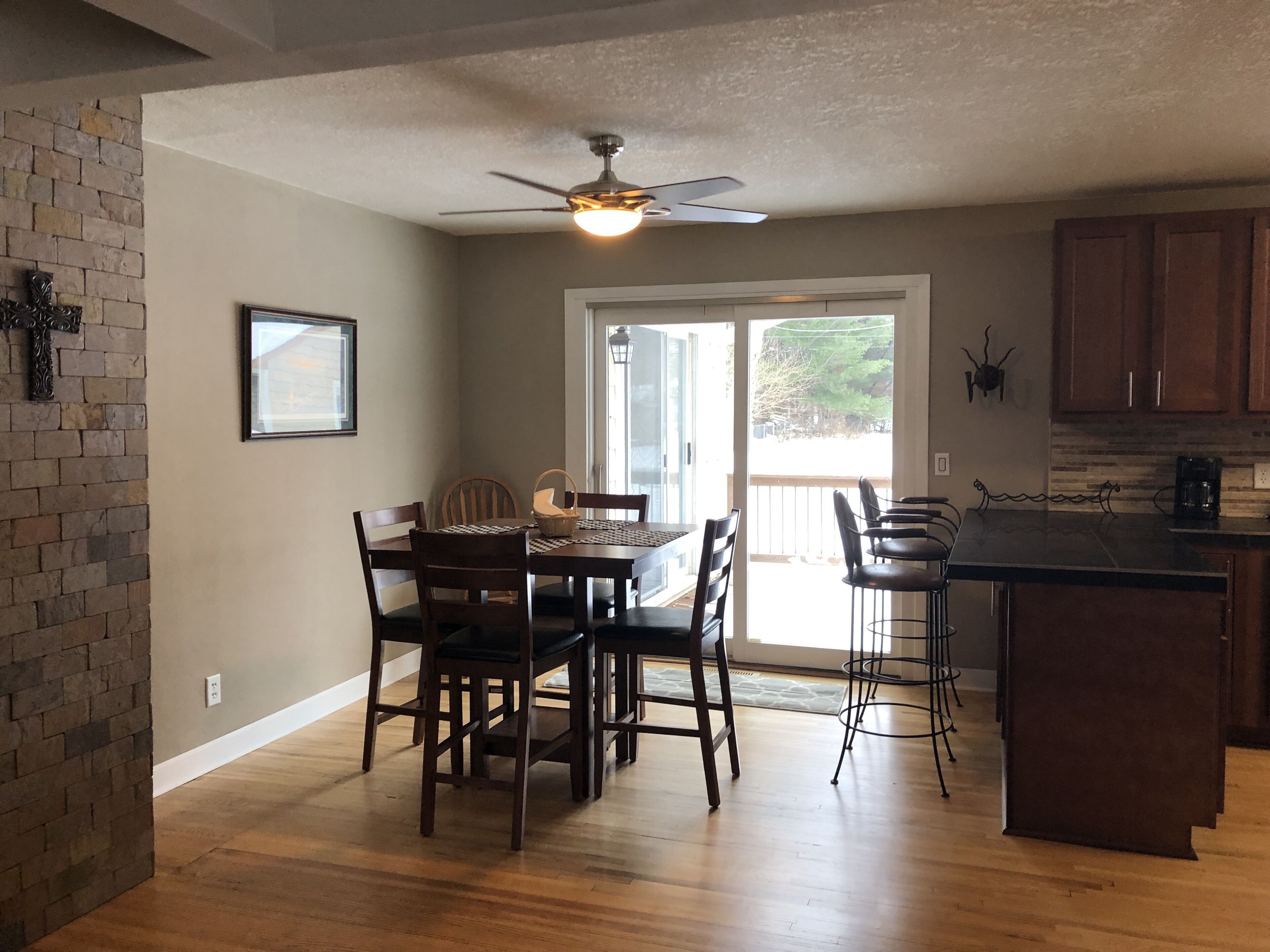 Un coin repas avec une table et des chaises en bois, un ventilateur de plafond et une vue sur une terrasse extérieure, accessible par une porte vitrée.
