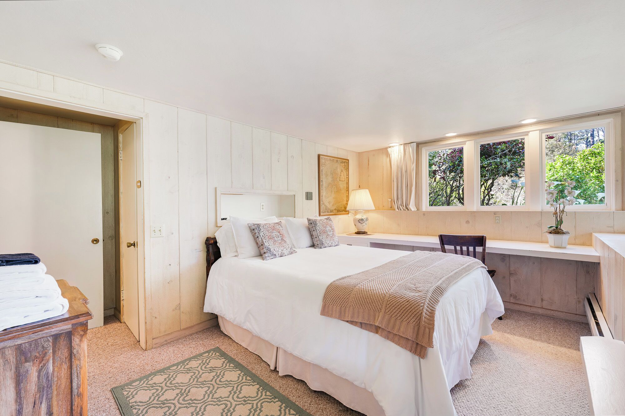 Un dormitorio con cama, mesita de noche, lámpara, ventana y vistas al verdor exterior.