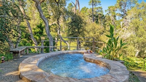 Un bain à remous circulaire, entouré d’une terrasse en pierre, avec une clôture en bois et une végétation luxuriante en arrière-plan.