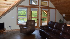 Un dormitorio tipo loft con techo alto, vigas de madera, un sofá de cuero marrón, un sillón de cuero y ventanas grandes que ofrecen vista a la vegetación exterior.